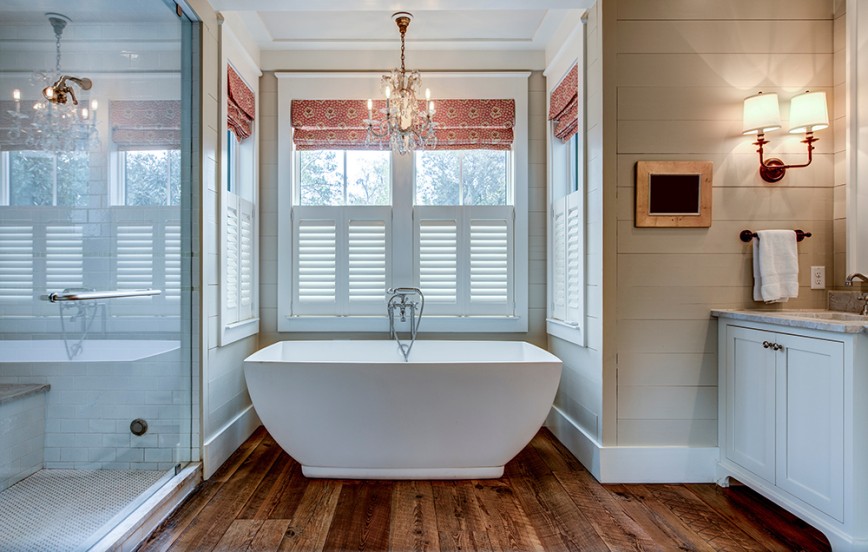 High end bathroom with large white bathtub and shiplap siding. Finished with chandelier lights and peach roman blinds and white shutters.